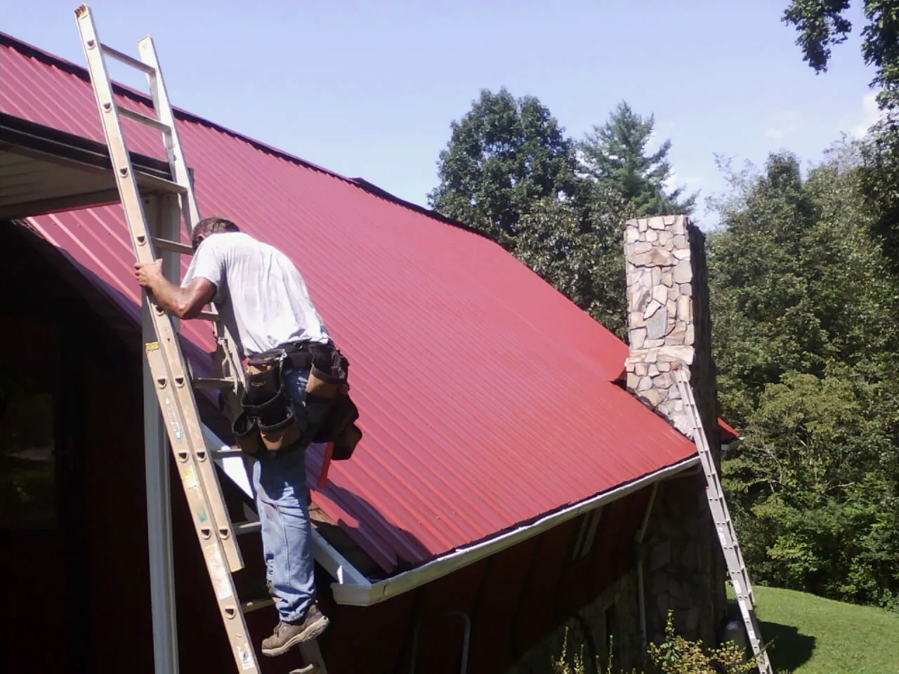Roofer on a ladder beside a red metal roof and stone chimney — Soffit & Fascia Repair by Roofing Brothers in Morrisville